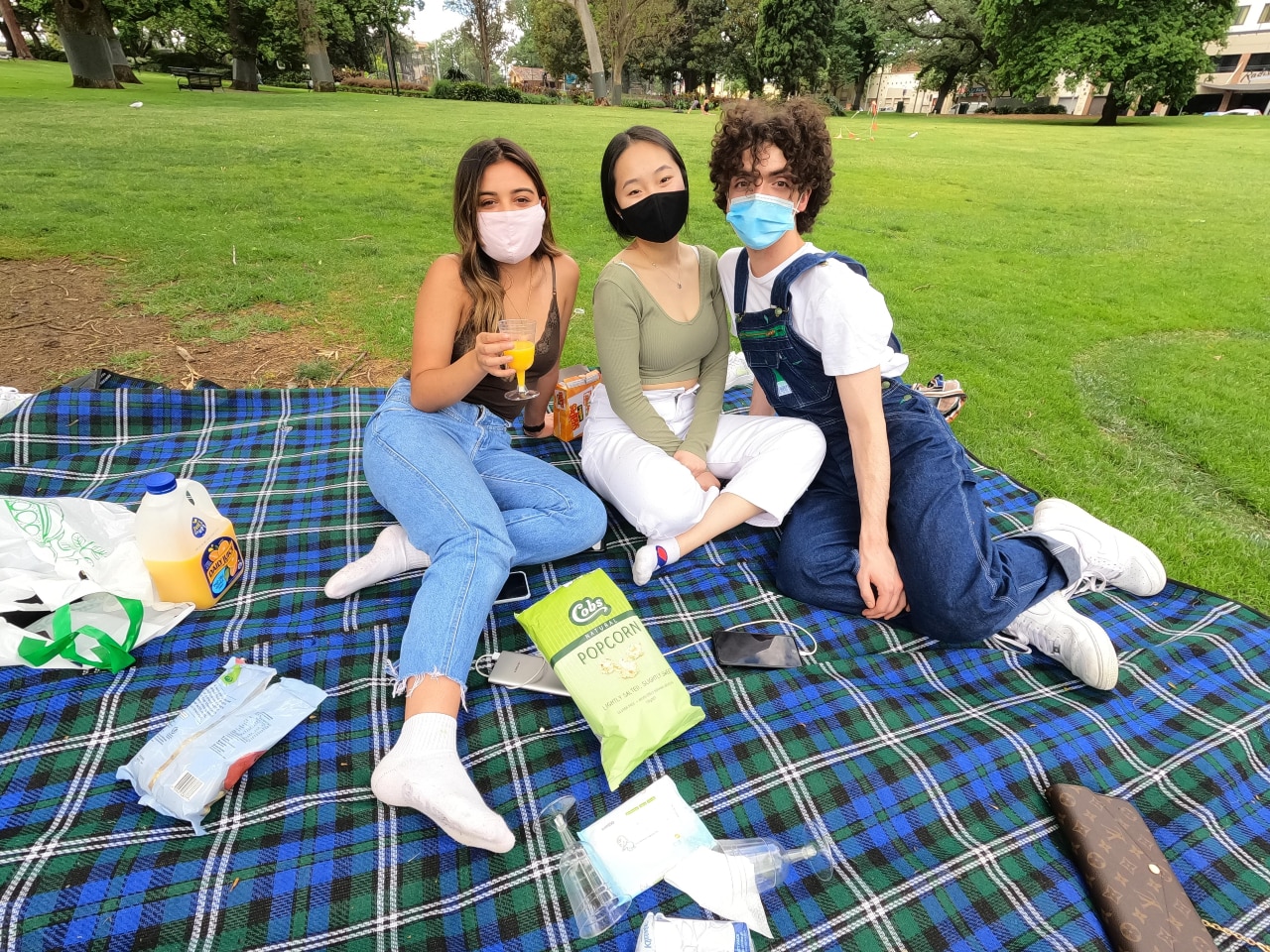 Three people, wearing face masks, sit on a picnic rug in a park.