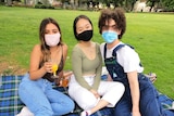 Three people, wearing face masks, sit on a picnic rug in a park.