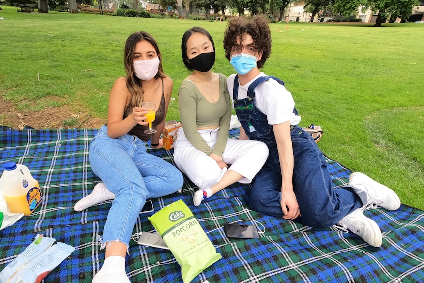 Three people, wearing face masks, sit on a picnic rug in a park.