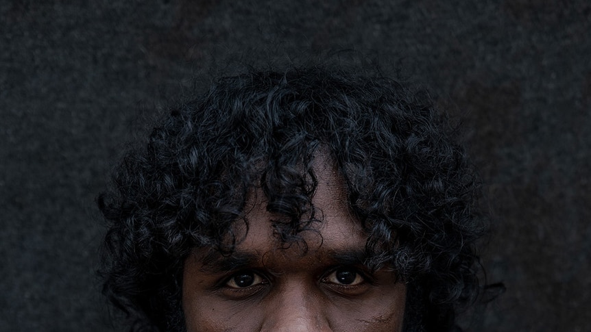 A shadowy, close-up portrait of Baykali Ganambarr in costume as an Indigenous tracker.