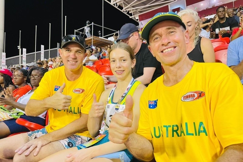 Two smiling men in bright "Australia" shirts sit either side of a young girl in stadium seats, giving the thumbs up.