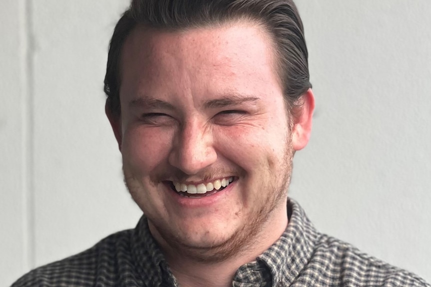 Photo of man smiling widely. He has brown slicked back hair and a checkered shirt.