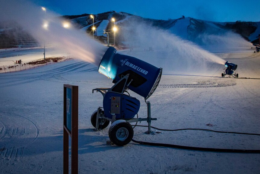 A snow machine blasts snow over ski slopes at night.
