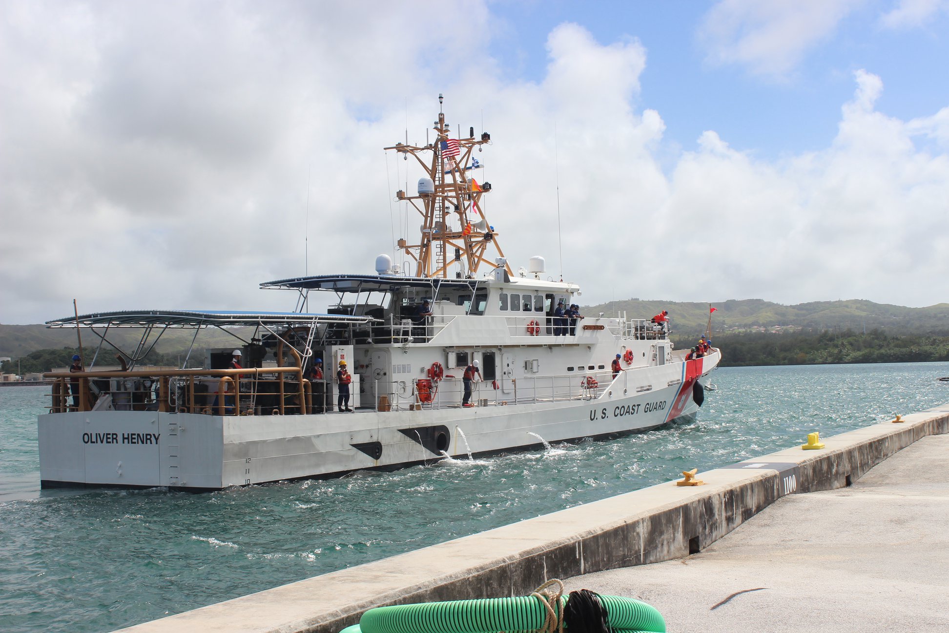 A white ship with a tall communications mast sits in bright blue water metres off a concret dock.