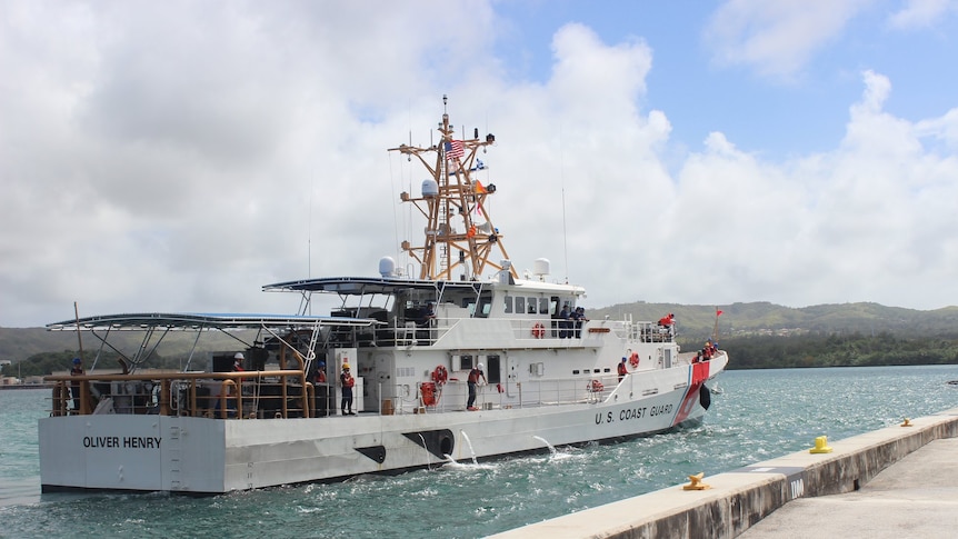 A white ship with a tall communications mast sits in bright blue water metres off a concret dock.