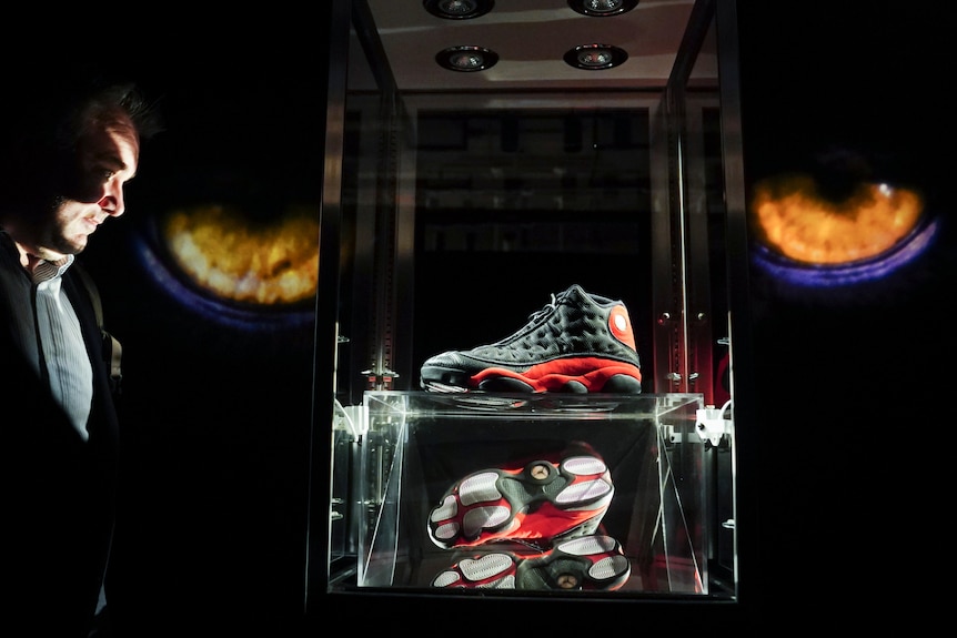 In a dark room a pair of black sneakers displayed behind glass, one upright, the other on its side. Left, a man looks at them.