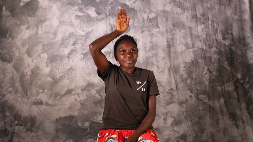 Indigenous woman sitting on a chair with plain backdrop performing sign language.