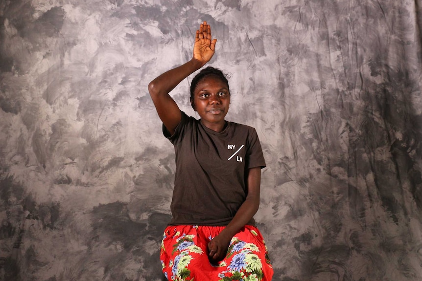 Indigenous woman sitting on a chair with plain backdrop performing sign language.