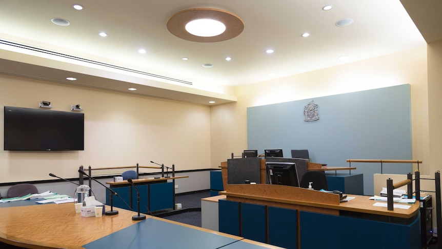 Benches equipped with monitors and microphones sit in a courtroom, with a large screen mounted on a wall.