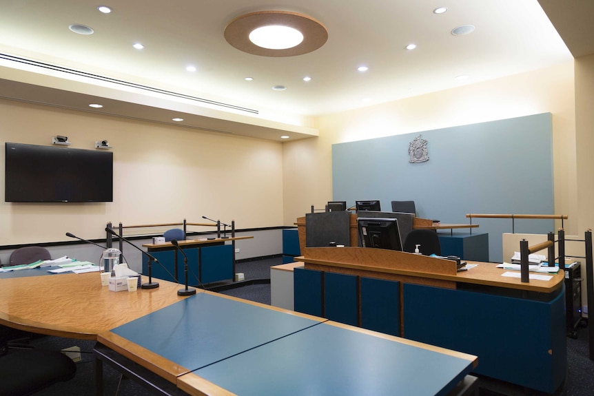 Benches equipped with monitors and microphones sit in a courtroom, with a large screen mounted on a wall.