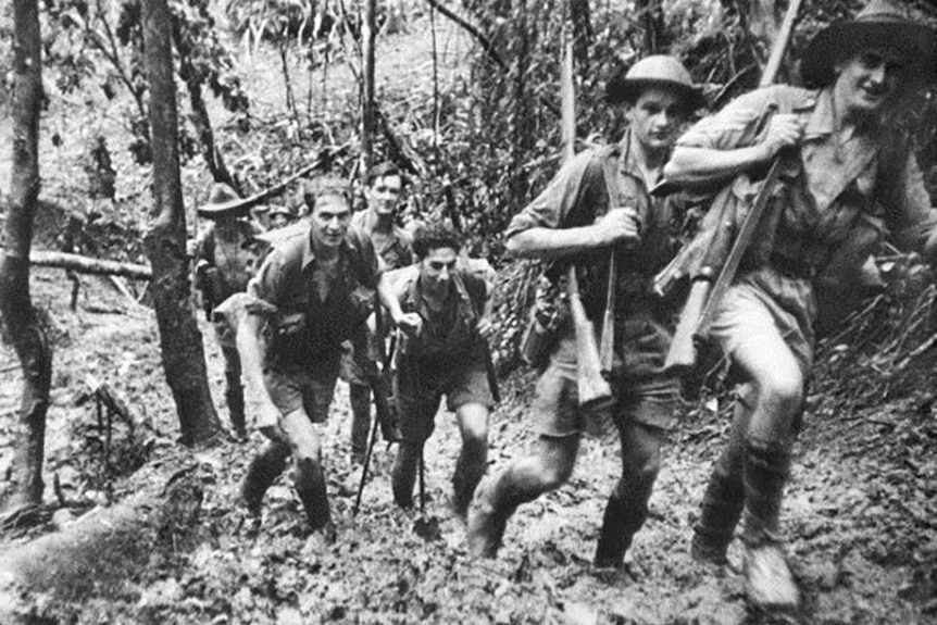 a black and white photograph of young men in uniform with rifles over their shoulders walking in the muddy  rainforest
