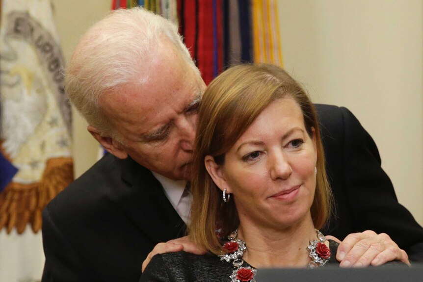 Joe Biden standing behind Stephanie Carter with his hand on her shoulders and mouth close to her ear