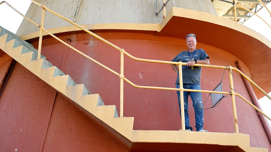 A man in painters jeans and a t-shirt stands on a yellow staircase at the base of a big dish.