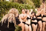 A woman with long blonde hair, wearing a black t-shirt, takes a photo with a DSLR camera of women smiling in black underwear