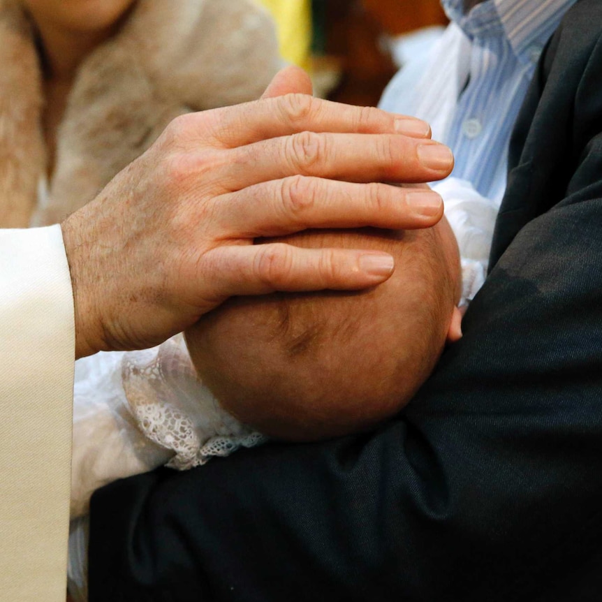 A priest's hand rests on a baby's head during a baptism.