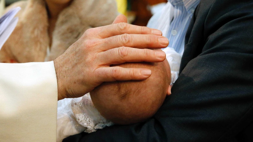 A priest's hand rests on a baby's head during a baptism.
