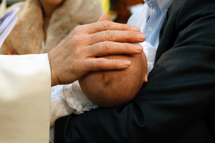 A priest's hand rests on a baby's head during a baptism.