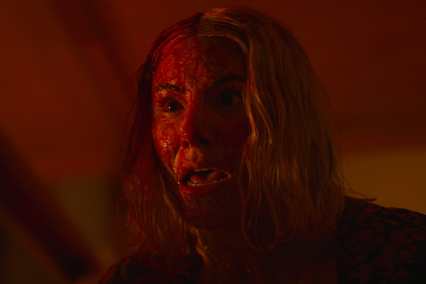 White woman with short blonde hair stands in darkened room, covered in blood and looking terrified.