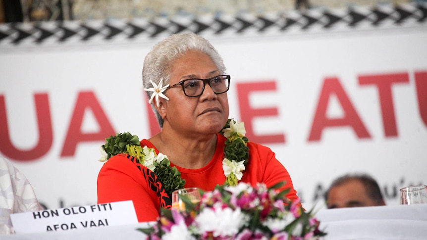 Woman wearing glasses and floral necklace with a flower behind right ear looks to the right of the frame.