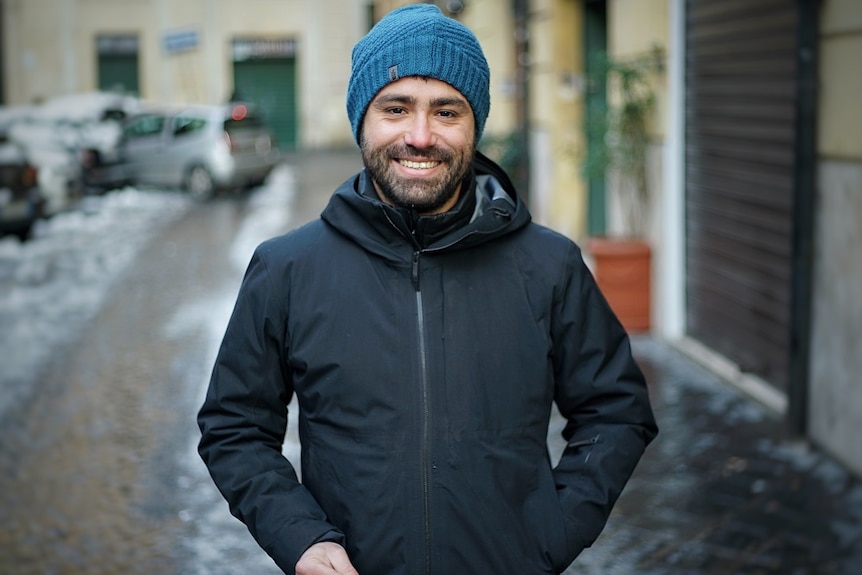 A young man wearing a beanie smiles in an Italian street.