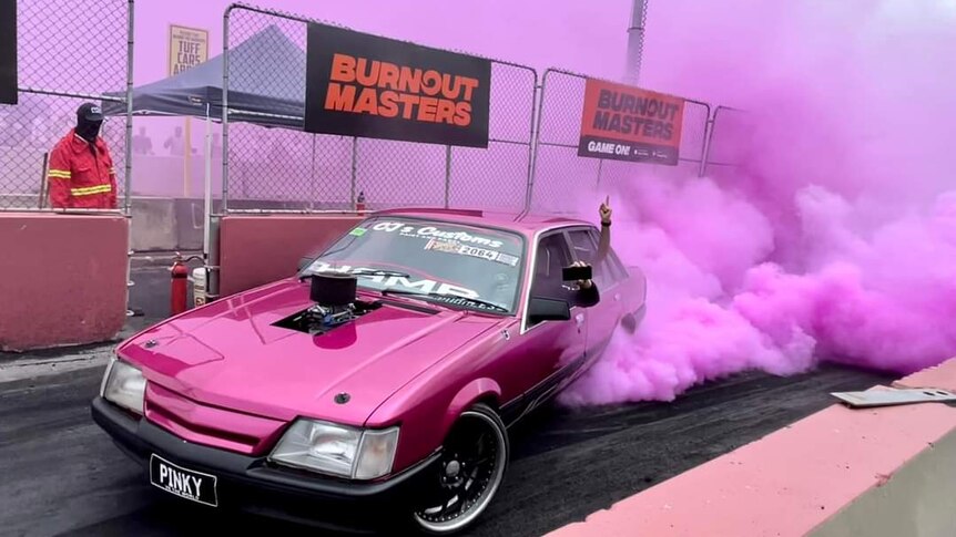 Bright pink smoke shoots from the tyres of a pink Holden Commodore burnout car with the number plate saying PINKY