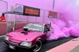 Bright pink smoke shoots from the tyres of a pink Holden Commodore burnout car with the number plate saying PINKY