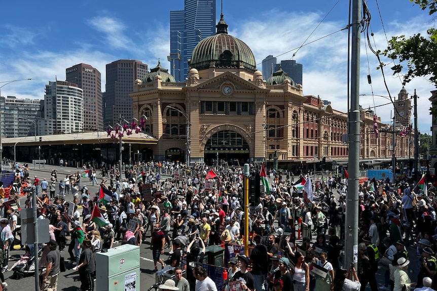 A crowd of people walks past Flinders Street Station in Melbourne on a sunny day as part of a protest.