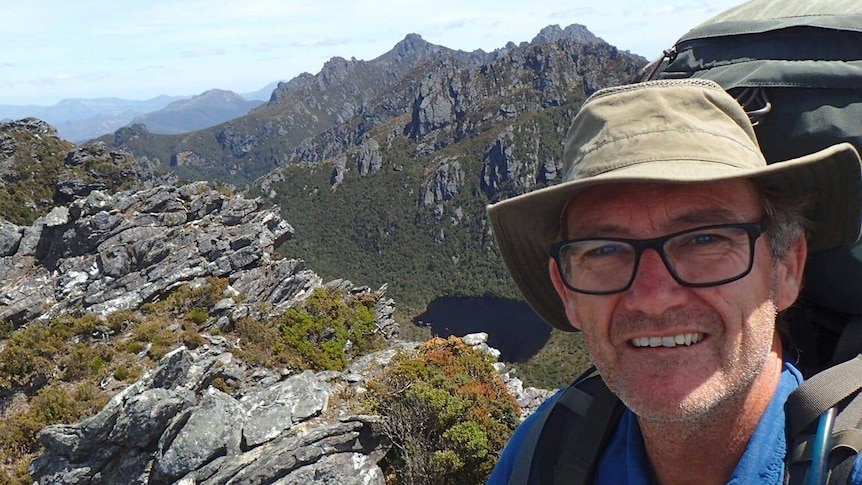 Neil Parker is wearing a hiking backpack and a hat, posing in front of a mountain view