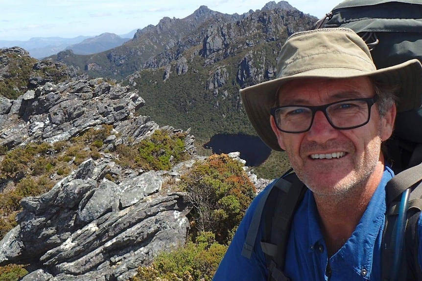 Neil Parker is wearing a hiking backpack and a hat, posing in front of a mountain view