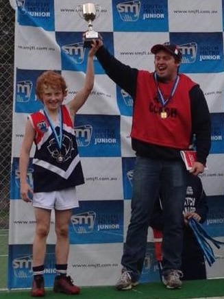 A father and son hold up a junior Australian rules trophy.