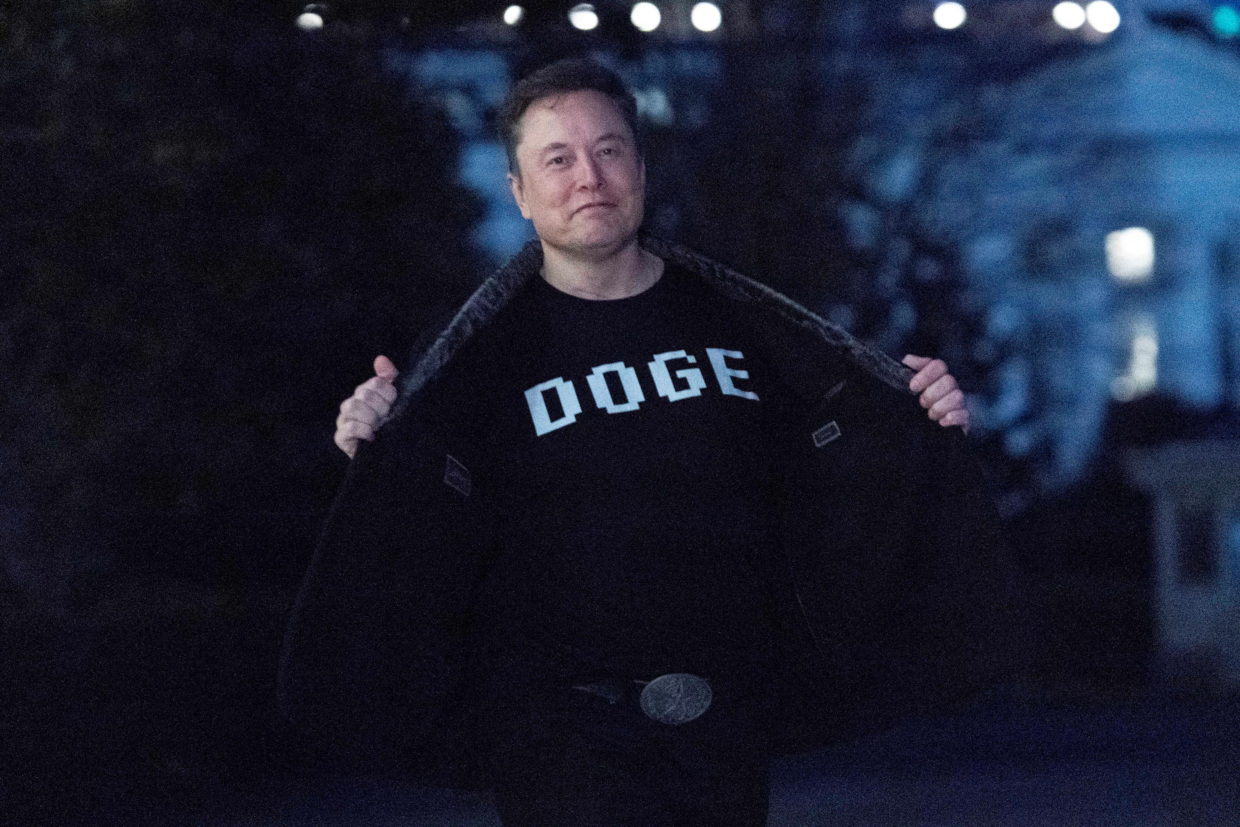 A man wearing a black shirt with a white logo that says "DOGE" shows it off to the camera smiling