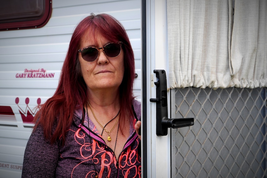 An older woman with dark red hair, wearing sunglasses, stands in front of a caravan.