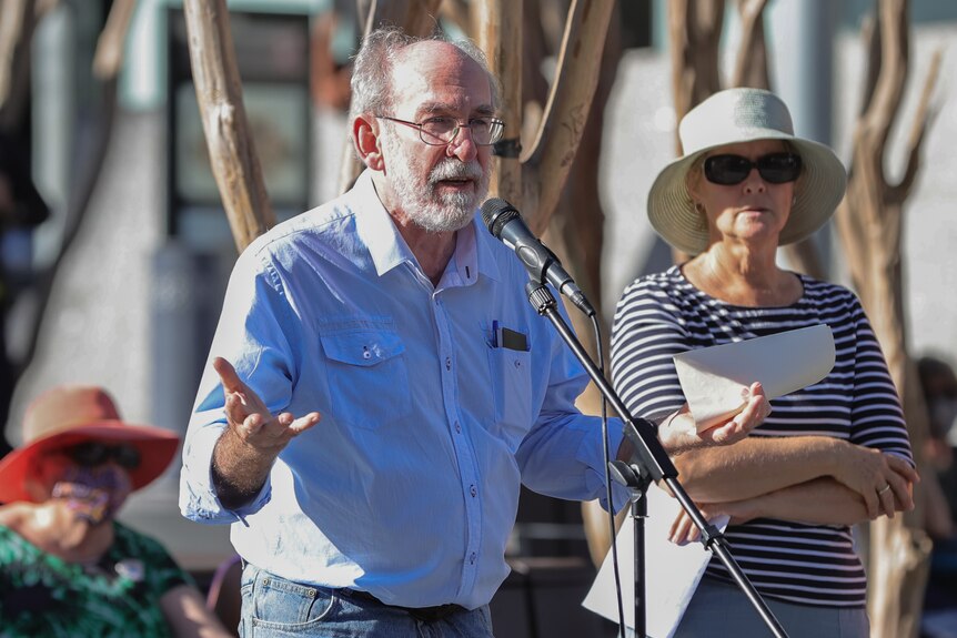 an older man wearing glasses addressing a crowd and talking into a microphone at a rally