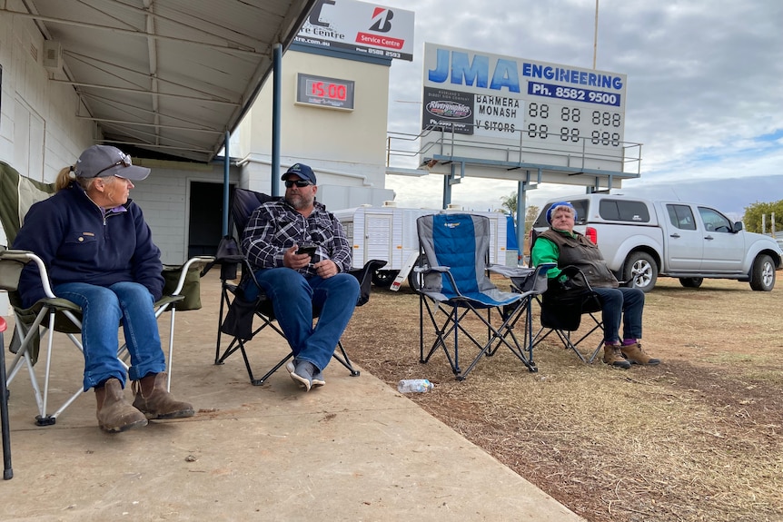 Three people sitting on camping chairs with a shed and scoreboard in background.