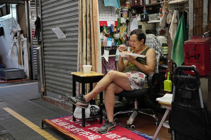 A man sits in a chair and eats out of a white polystyrene container with chopsticks.