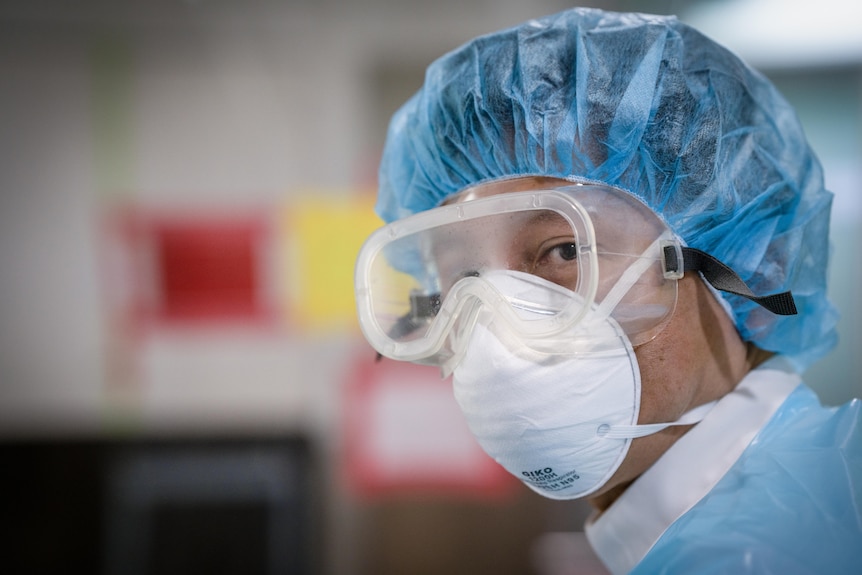 A Japanese man in a mask, fogged goggles and a blue shower cap looks at the camera