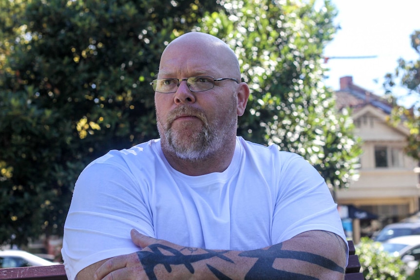 A bald man wearing glasses and a white T-shirt stands with his arms crossed, one of which is decorated with black tattoos.