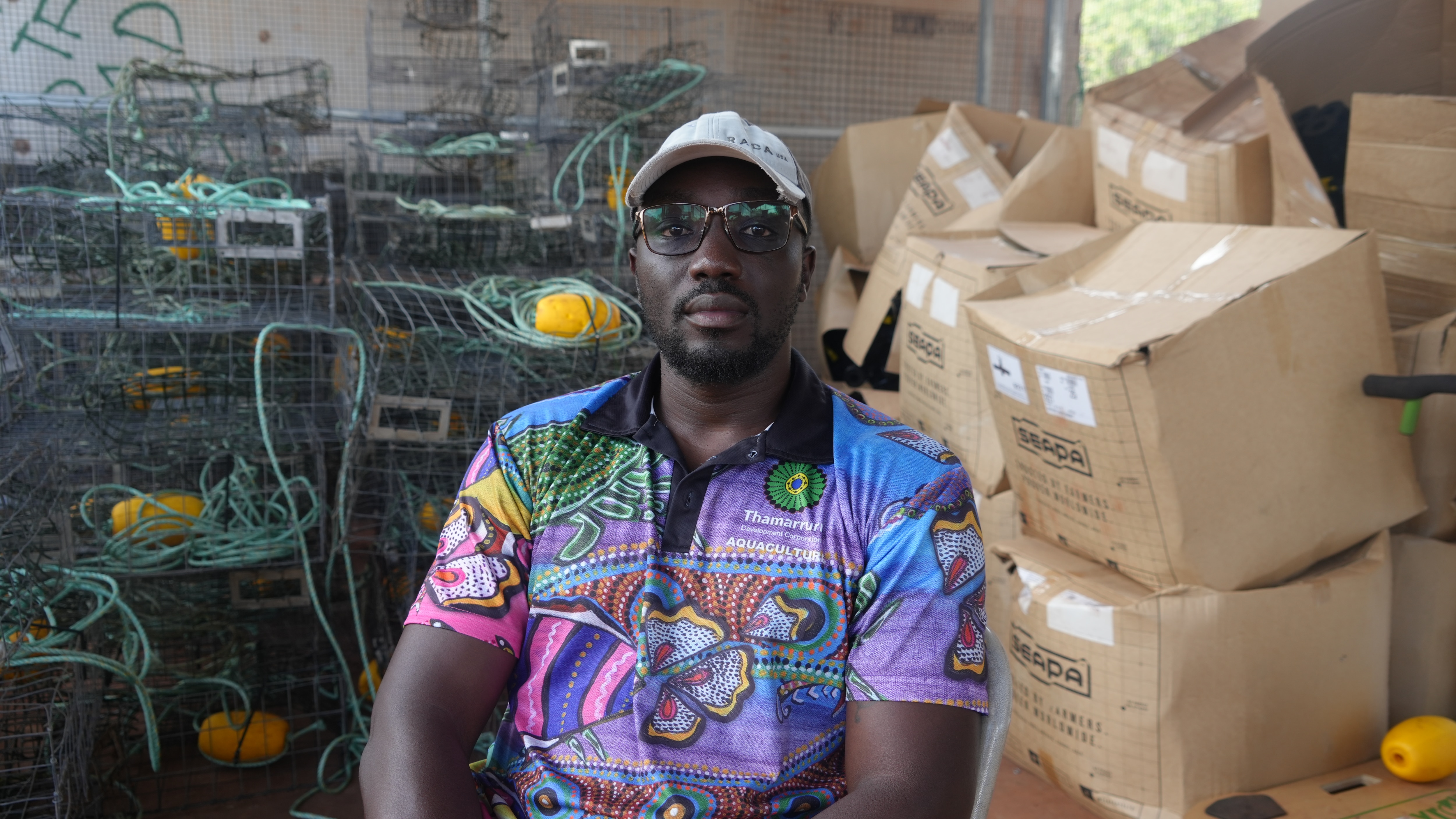 A Black man wearing a colourful shirt. There are several crab pots in the background and large cardboard boxes.