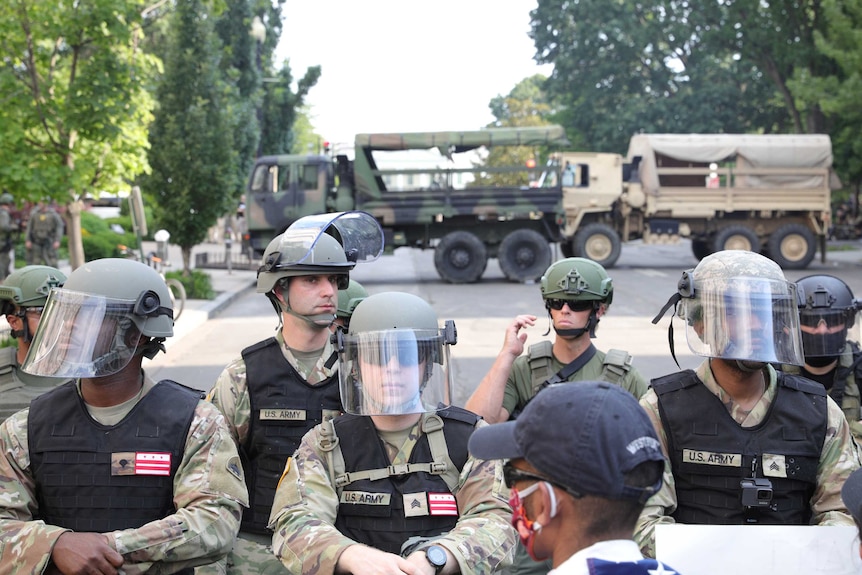 Men dressed in military fatigues and wearing helmets stand in front of protests