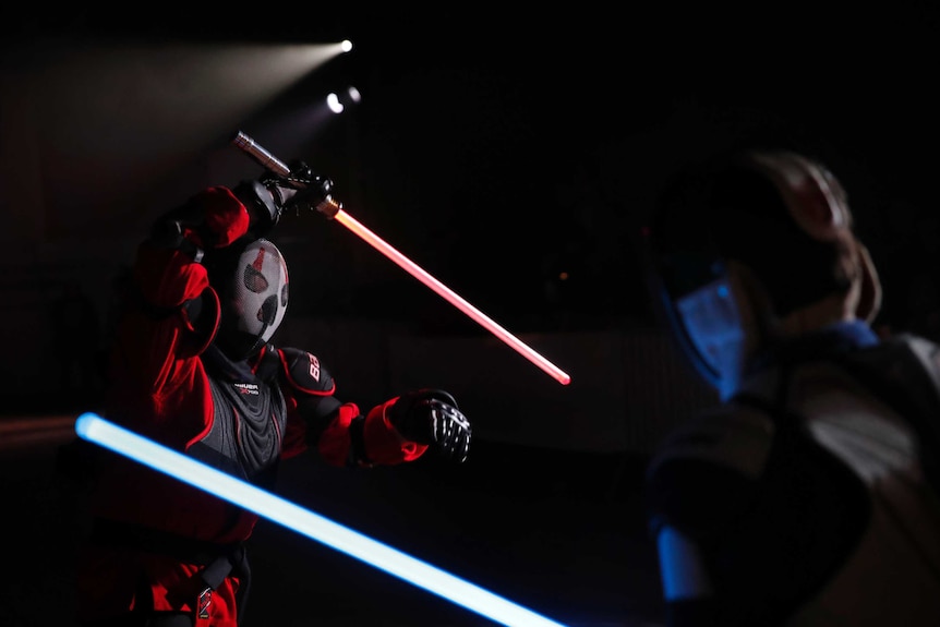 A masked man brandishes a red lightsaber over his head while his opponent defends himself.