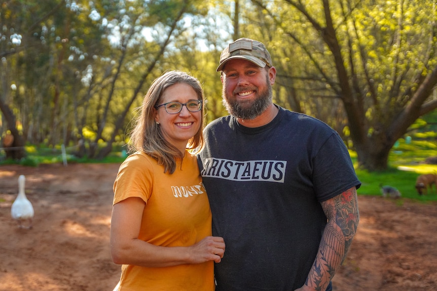 A man stands with his arm around a woman outside next to trees at sunset, both smiling.