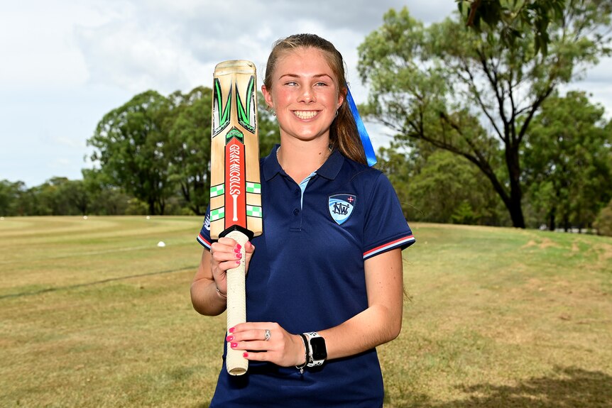 Courtney Webeck smiles while holding a cricket bat over her shoulder.