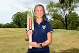 Courtney Webeck smiles while holding a cricket bat over her shoulder.