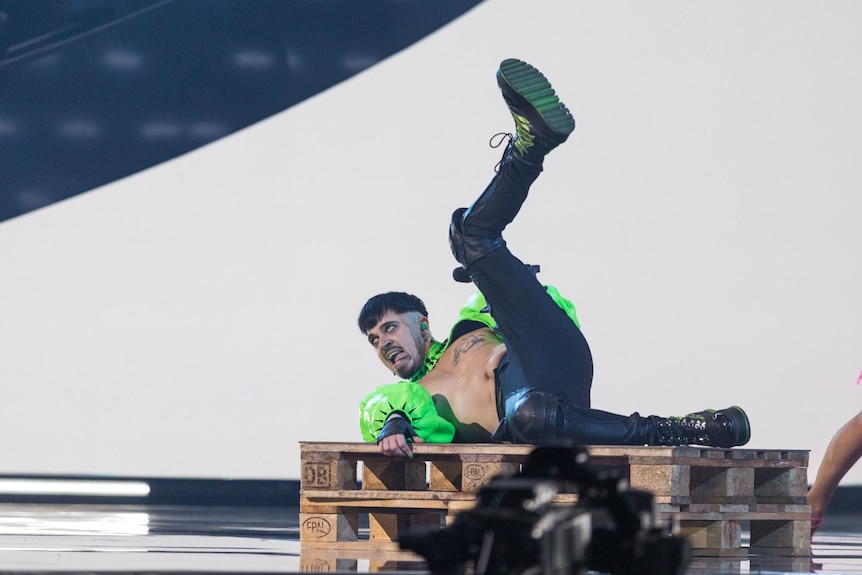 A man on stage in a green top, chest exposed, laying down with leg in air