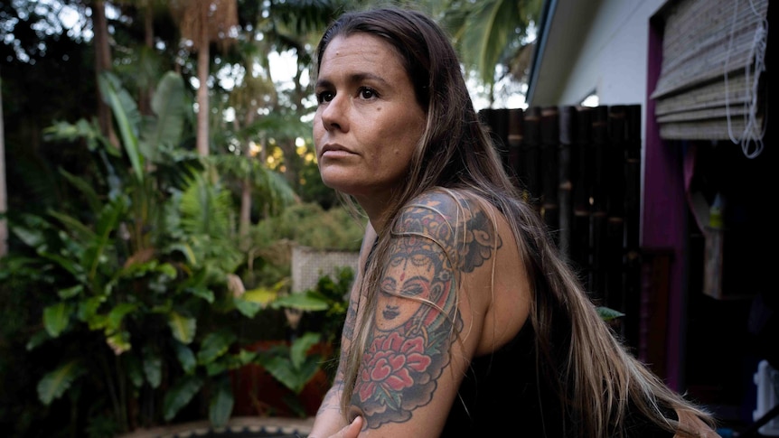 A woman with colourful tattoos on her arms leans on a railing and stares into the distance. A pool & palm trees are behind her.
