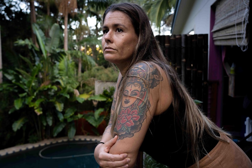 A woman with colourful tattoos on her arms leans on a railing and stares into the distance. A pool & palm trees are behind her.