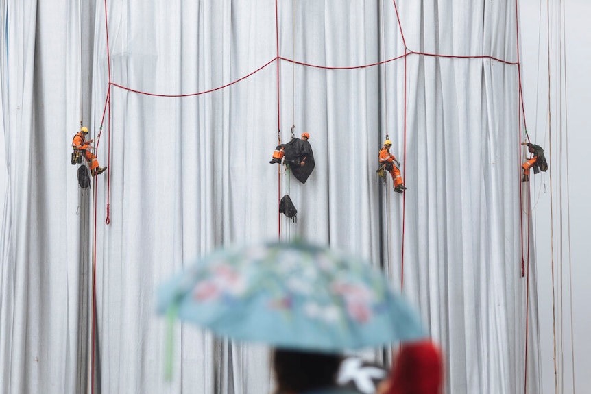 Workers hanging from ropes against a veil of fabric.