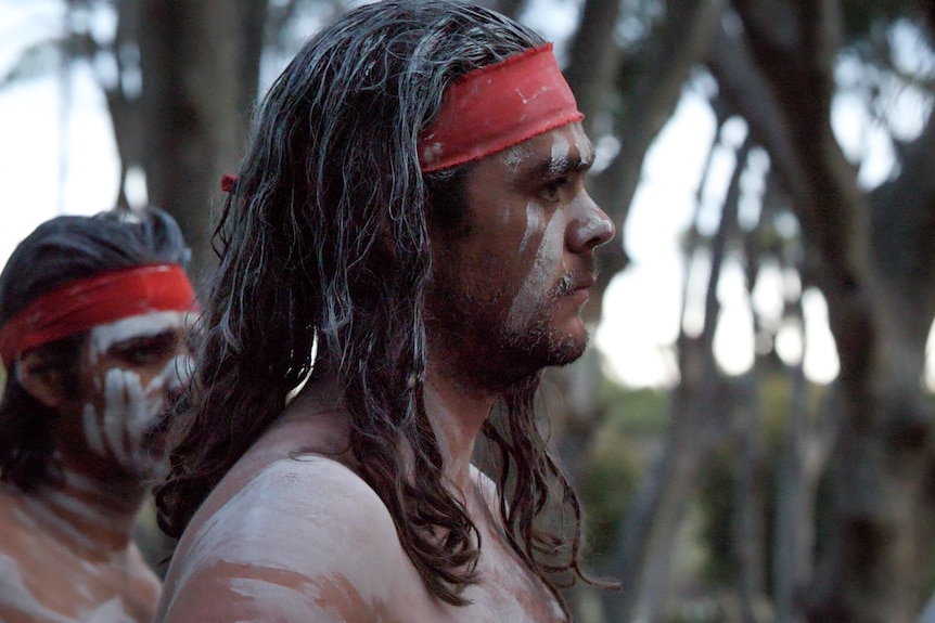Two men in traditional ochre and red headband standing in forest watching a dance