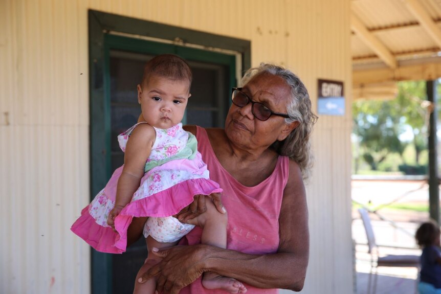 Indigenous woman with glasses and grey hair stands with baby wearing pink frilly dress on her hip