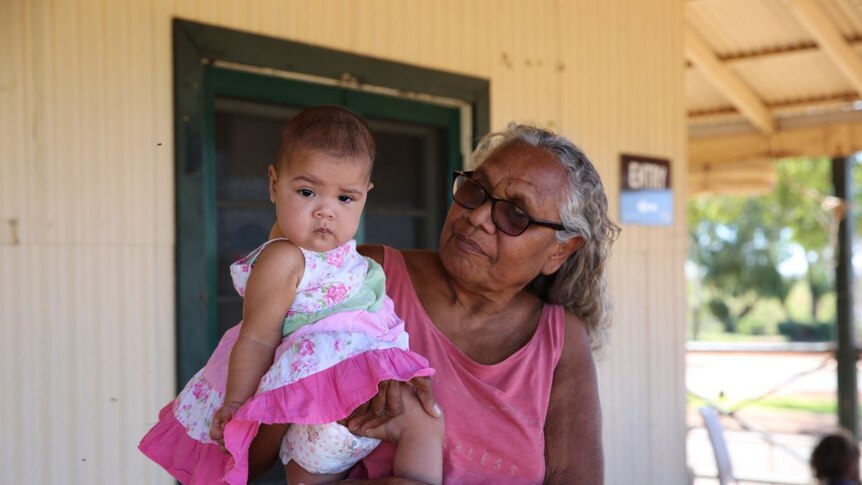 Indigenous woman with glasses and grey hair stands with baby wearing pink frilly dress on her hip